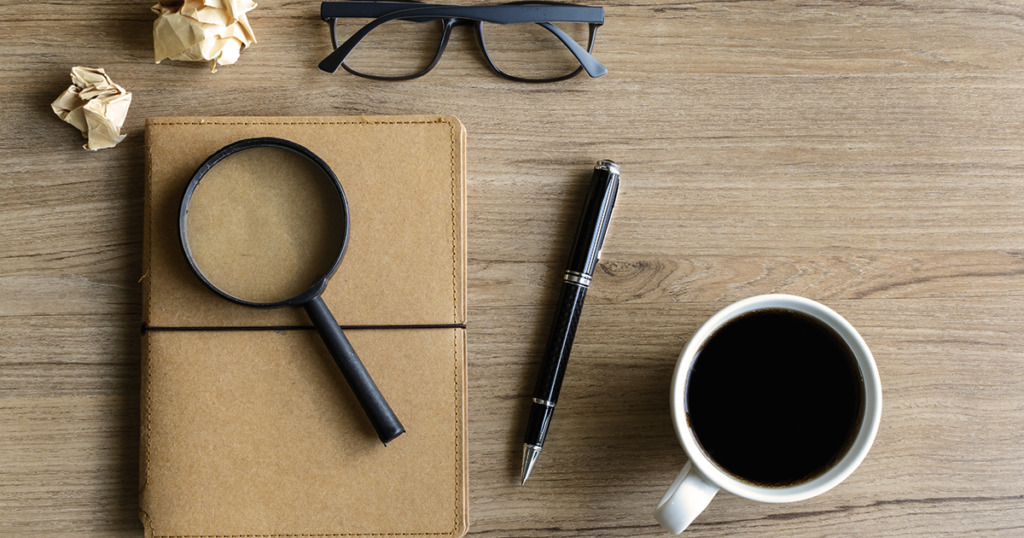 Leather journal, magnifying glass, fountain pen, and coffee cup arranged on a wooden desk
